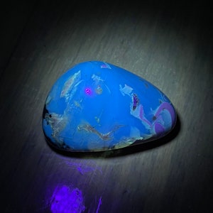 May include: A blue, white, and purple stone with a speckled pattern under a UV light. The stone glows a bright blue color.