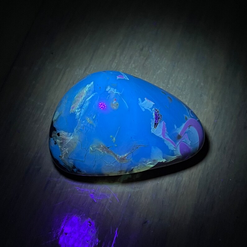 May include: A blue, white, and purple stone with a speckled pattern under a UV light. The stone glows a bright blue color.