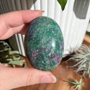 Puede incluir: Gema de forma ovalada con superficie pulida. La piedra presenta una base verde moteada con pequeñas inclusiones rojas vibrantes. La gema se sostiene en una mano, mostrando su textura suave y belleza natural.