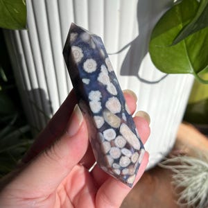 May include: A purple and white agate crystal point with a unique, speckled pattern. The crystal is smooth and polished, with a pointed tip.