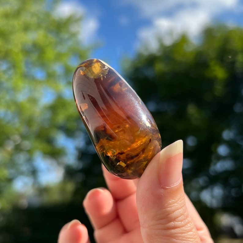 May include: A smooth, polished amber stone with a warm, honey-brown color and flecks of gold. The stone is oval-shaped and has a glossy finish.