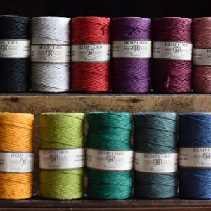 May include: A selection of colourful hemp cord spools in shades like black, white, red, purple, and pink. The spools are stacked on a wooden surface, with labels visible. The image showcases a variety of craft supplies for creative projects.