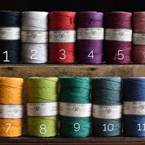 May include: A collection of hemp cord spools in a range of colours, such as black, white, red, purple, and pink. Each spool is labelled with a number and the words "Hemp Cord". The cords are stacked on a wooden surface.