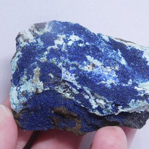 May include: A rough, natural azurite mineral specimen. The stone features a deep blue color with areas of lighter blue and white, creating a striking contrast. The rock has a textured surface and is held in a hand.
