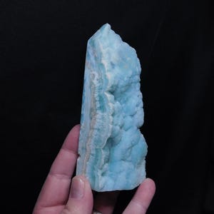 May include: A light blue aragonite crystal with a rough, textured surface. The crystal has a layered appearance with areas of white and light blue. The crystal is held against a black background.