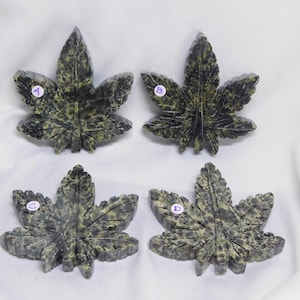 May include: Four carved stone leaf sculptures with a cannabis leaf design. The leaves are a mottled green and gray color, with detailed veining. Each leaf is labeled with a letter A, B, C, or D on a small white sticker.