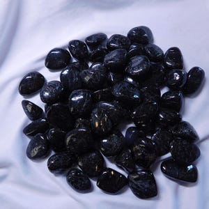 May include: A pile of black tumbled stones with a shimmery, iridescent finish. The stones are smooth and polished, and they are arranged in a loose pile on a white background.