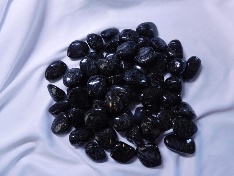 May include: A pile of black tumbled stones with a shimmery, iridescent finish. The stones are smooth and polished, and they are arranged in a loose pile on a white background.