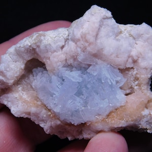 May include: A close-up of a mineral specimen featuring a light pink matrix with a cluster of pale blue crystals in the center. The mineral has a rough, textured surface and is set against a dark background.