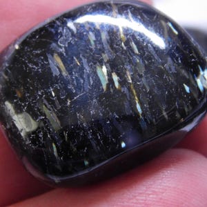 May include: A black gemstone with iridescent blue and gold streaks. The gemstone is smooth and polished, and has a rounded shape.