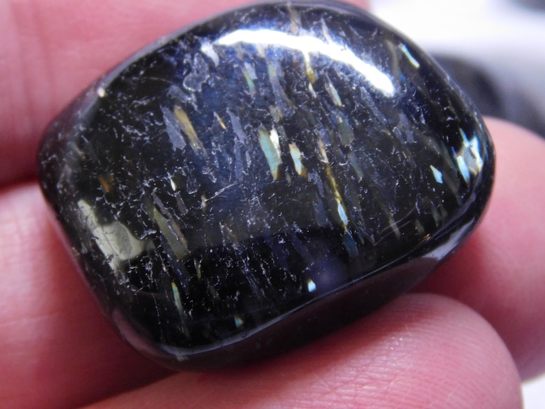 May include: A black gemstone with iridescent blue and gold streaks. The gemstone is smooth and polished, and has a rounded shape.