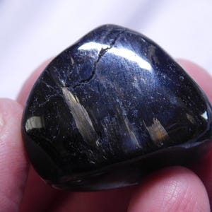 May include: A smooth, black gemstone with iridescent blue and gold streaks. The stone is polished and has a rounded shape.