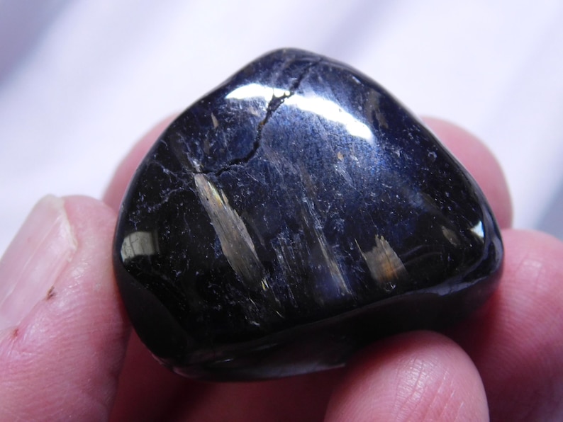 May include: A smooth, black gemstone with iridescent blue and gold streaks. The stone is polished and has a rounded shape.