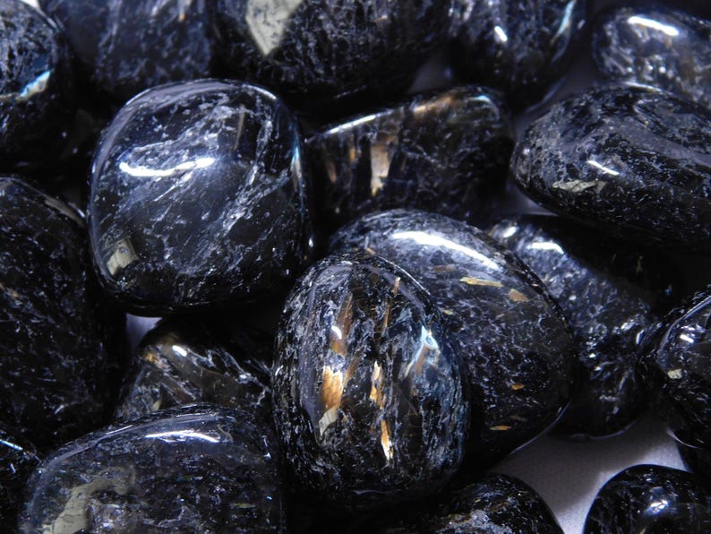 May include: A collection of black obsidian tumbled stones with a shimmering, iridescent effect. The stones are smooth and polished, with a variety of shapes and sizes.