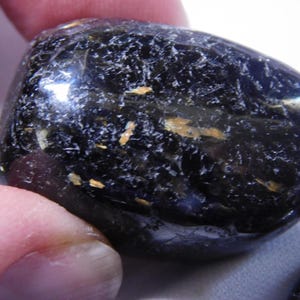May include: A smooth, black gemstone with gold flecks. The stone is polished and has a rounded shape.