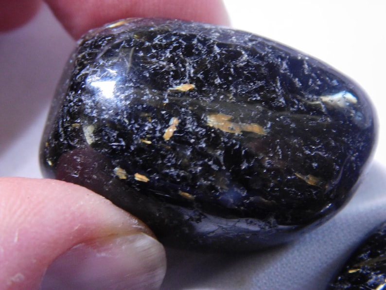 May include: A smooth, black gemstone with gold flecks. The stone is polished and has a rounded shape.
