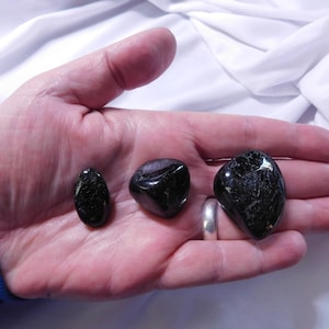 May include: Three black tumbled stones, two are oval shaped and one is heart shaped, are displayed on a hand.