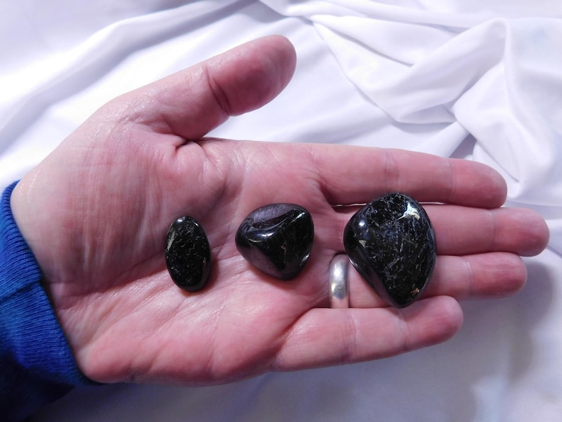 May include: Three black tumbled stones, two are oval shaped and one is heart shaped, are displayed on a hand.