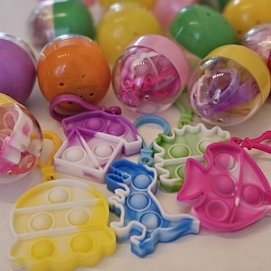 May include: A collection of colorful plastic toy eggs and pop-it keychains. The eggs are in various colors, some transparent, and the keychains are shaped like a dinosaur, a fish, and other shapes. The pop-its are in shades of blue, pink, yellow, and green.