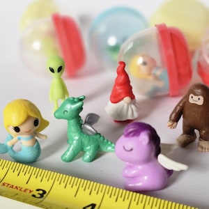 May include: A collection of small, colorful figurines including a mermaid, dragon, unicorn, alien, gnome, and Bigfoot. Several clear plastic capsules with red and yellow tops are in the background. A yellow tape measure is in the foreground.