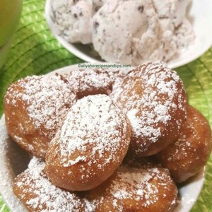 May include: A white bowl filled with golden-brown beignets, generously dusted with icing sugar. In the background, a separate white bowl holds scoops of cookies and cream ice cream. The image is set against a green background.