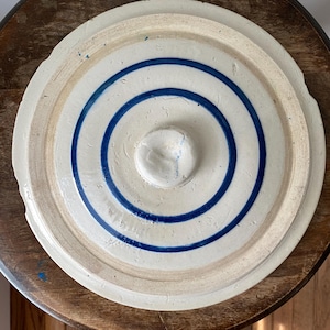 May include: A circular, off-white stoneware lid featuring a central knob and two concentric blue rings painted on the surface. The lid has a stepped edge design with a slightly uneven, textured finish and visible signs of wear along the rim.