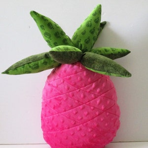 May include: A pink pineapple-shaped pillow with a green leafy top. The pillow is made of a soft, textured fabric and has a quilted design.