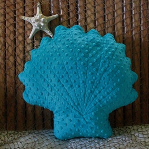 Op de afbeelding: Een turquoiseblauw pluche kussen in de vorm van een schelp met een textuur. Het kussen is gevormd als een schelp en heeft een driedimensionaal effect.