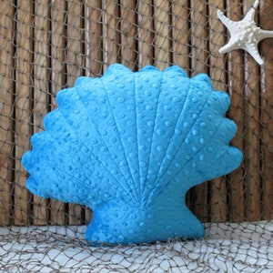 Op de afbeelding: Een blauw pluche kussen in de vorm van een schelp met een textuur. Het kussen ligt op een bruin en wit net.