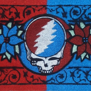 May include: A red and blue doormat with a black and white Grateful Dead logo in the center. The logo is surrounded by two black flowers, one on each side. The doormat has a black border with a floral design.