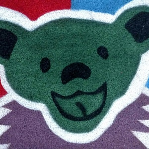 May include: A green bear with a white outline and a purple background. The bear has a big smile and a long tongue sticking out. The bear is on a red, white, and blue background.