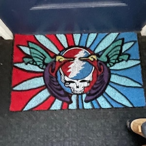 May include: A doormat with a red, white, and blue design featuring a Grateful Dead Steal Your Face logo with two hummingbirds on either side.