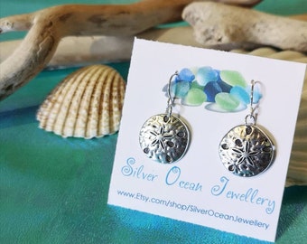 May include: Silver dangle earrings featuring a sand dollar design. The earrings are on a white card with the text "Silver Ocean Jewellery" and a website address.
