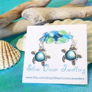 Può includere: Orecchini pendenti in argento con un ciondolo a forma di tartaruga blu iridescente. Gli orecchini sono esposti su una carta bianca con il testo "Silver Ocean Jewellery" e un indirizzo del sito web.