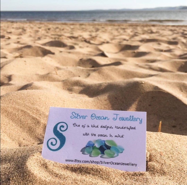 May include: A white card with blue text and a blue swirl design reads "Silver Ocean Jewellery" and "One of a kind designs, handcrafted with the ocean in mind." The card also includes the website address "www.Etsy.com/shop/SilverOceanJewellery". The card is lying on a sandy beach.