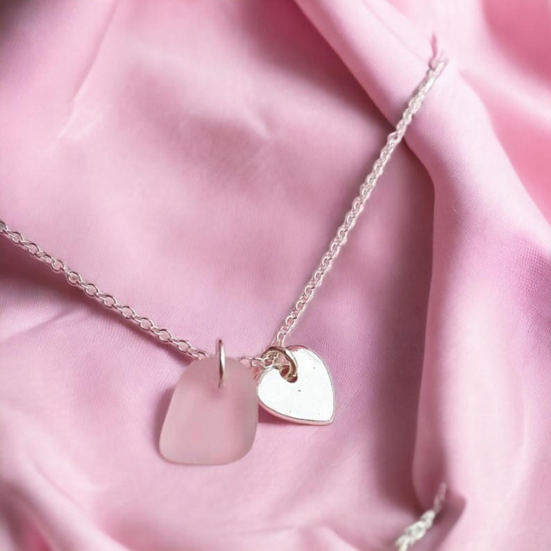 Pode incluir: Um colar de corrente de prata com um pingente de vidro fosco e um berloque de cora&ccedil;&atilde;o de prata. O colar &eacute; apresentado em um fundo de tecido rosa.