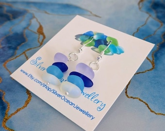 Puede incluir: Un par de pendientes colgantes de plata con piedras de vidrio de mar azul y morado. Los pendientes están en una tarjeta blanca con el texto "Silver Ocean Jewellery" y una dirección de sitio web.
