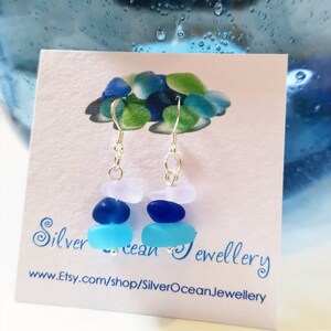 May include: A pair of silver dangle earrings with three sea glass beads in shades of blue and white. The earrings are on a white card with the text "Silver Ocean Jewellery" and a website address.