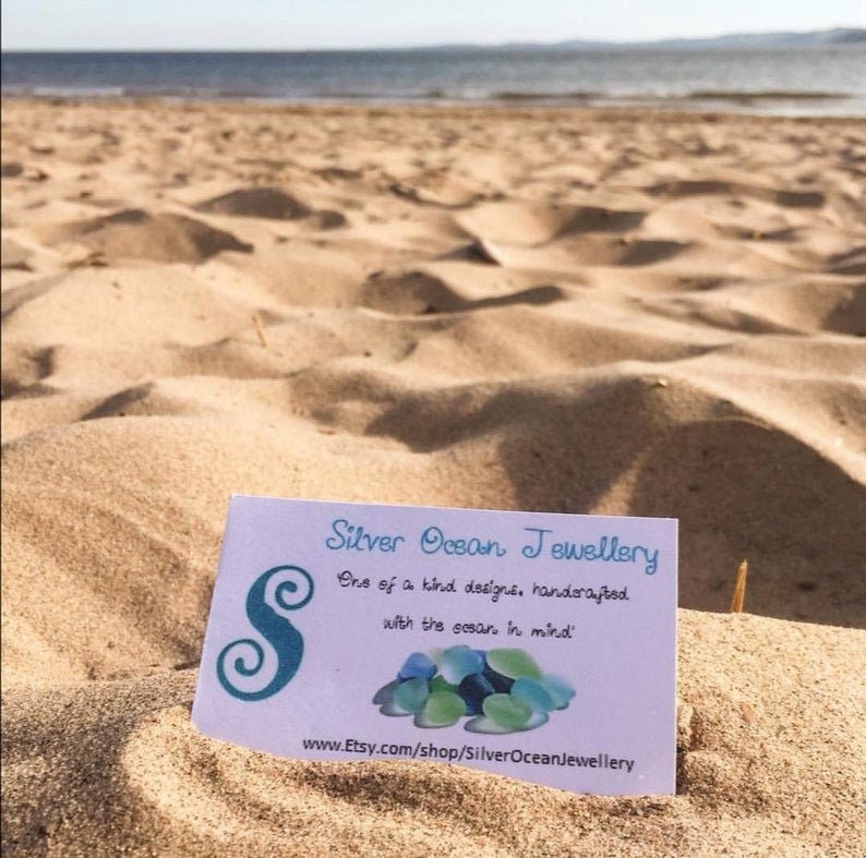 Pode incluir: Um cart&atilde;o de visita para Silver Ocean Jewellery, uma empresa que fabrica joias artesanais inspiradas no oceano. O cart&atilde;o apresenta um design de vidro do mar azul e verde e o texto "Designs &uacute;nicos, feitos &agrave; m&atilde;o com o oceano em mente." e "www.Etsy.com/shop/SilverOceanJewellery".