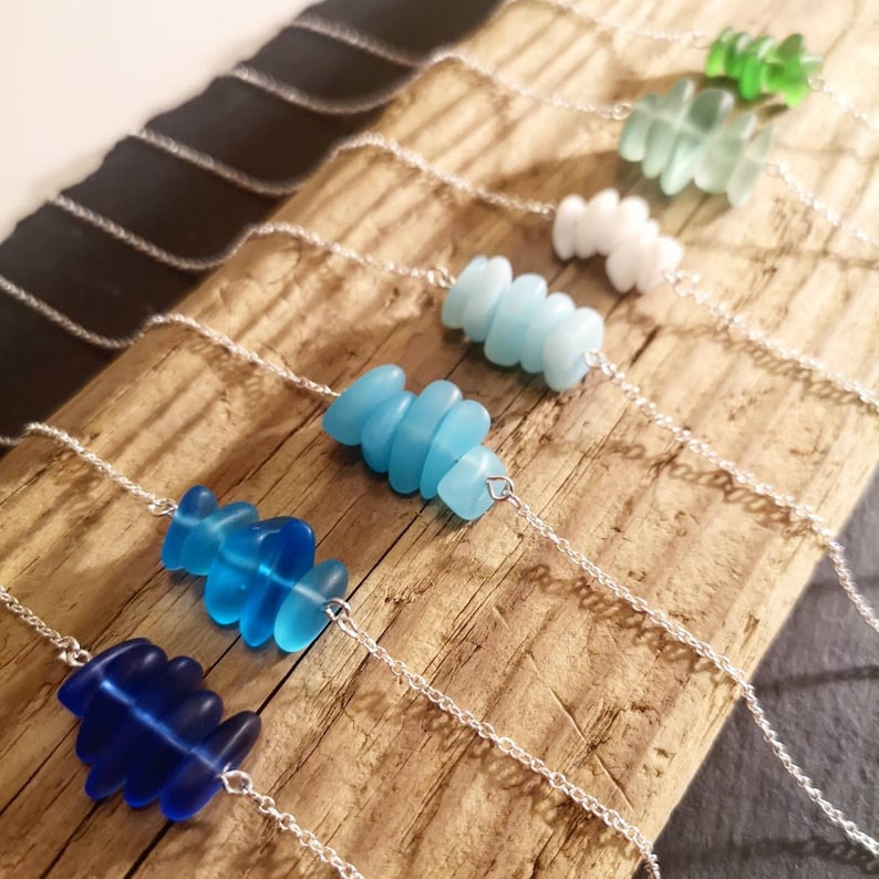 May include: A set of seven necklaces with graduated shades of blue sea glass, each necklace features a different number of sea glass pieces, the necklaces are displayed on a wooden surface.