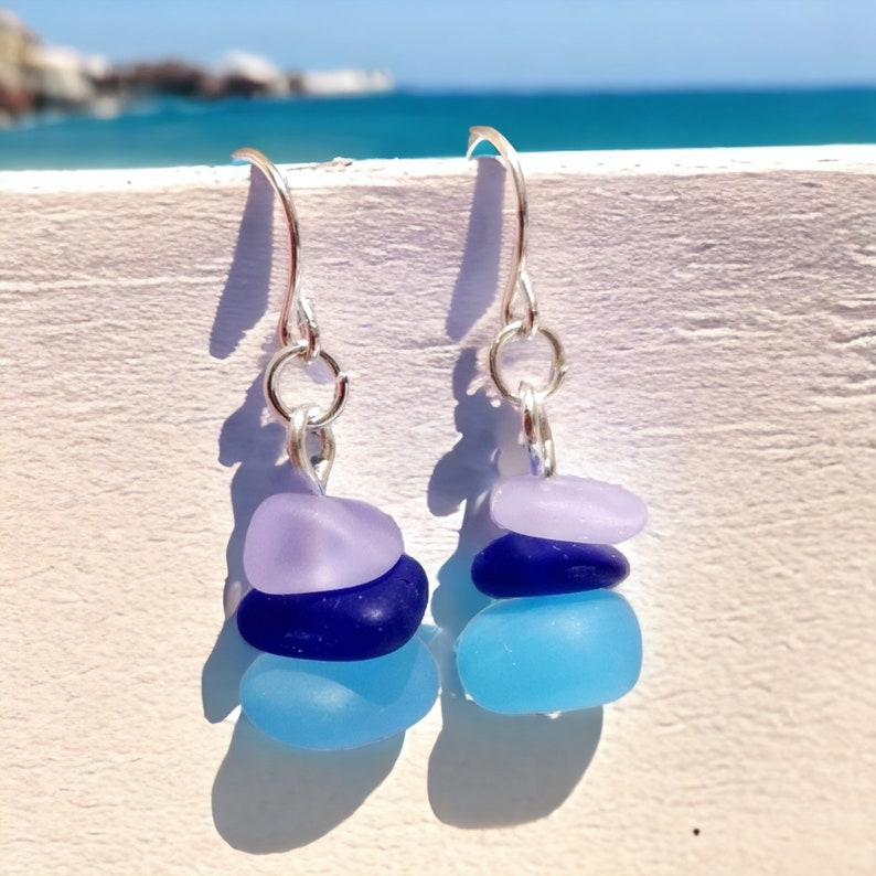 May include: A pair of silver dangle earrings with three stacked sea glass stones in shades of blue and purple.