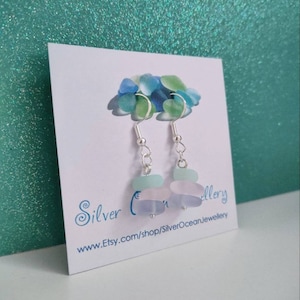 May include: A pair of silver dangle earrings with three layered sea glass beads in shades of blue and purple. The earrings are displayed on a white card with the text "Silver Ocean Jewellery" and a website address.
