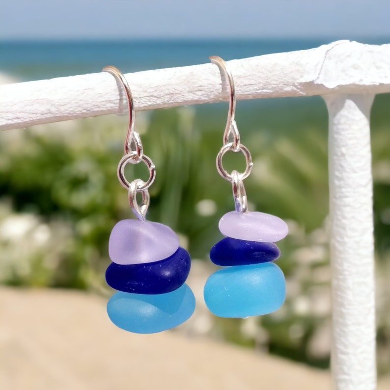 May include: A pair of silver dangle earrings with three stacked sea glass stones. The stones are layered from top to bottom in shades of lavender, dark blue, and light blue.