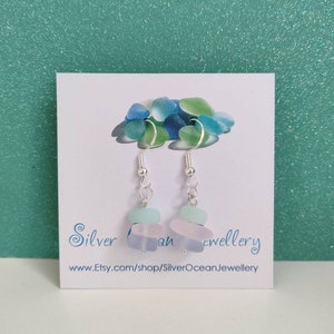 May include: Silver dangle earrings with three layered sea glass beads in shades of blue and white. The earrings are on a white card with the text "Silver Ocean Jewellery" and a website address.