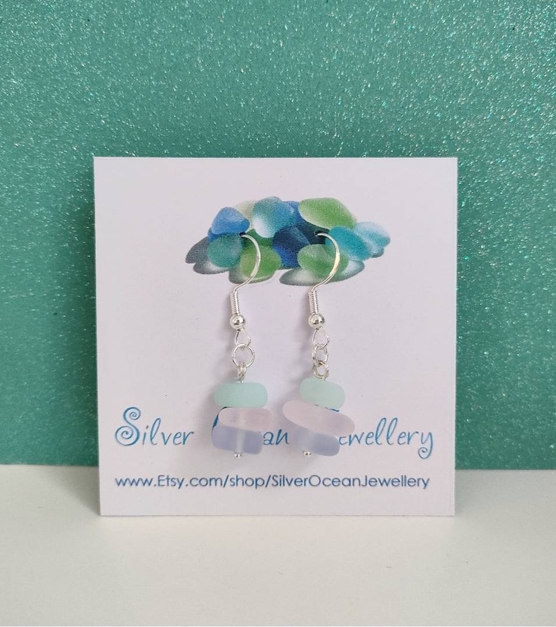 May include: Silver dangle earrings with three layered sea glass beads in shades of blue and white. The earrings are on a white card with the text "Silver Ocean Jewellery" and a website address.