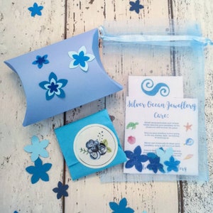 May include: A blue pillow box with blue paper flowers, a blue tissue paper envelope with a floral design, and a card with the text "Silver Ocean Jewellery Care:" and instructions for caring for jewelry.