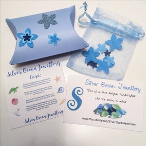 May include: A blue and white card with the text "Silver Ocean Jewellery" and care instructions for jewelry. The card is next to a small blue organza bag with blue and white flower cutouts. The bag is on a white surface.