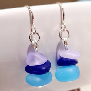 May include: Silver dangle earrings with three layered sea glass stones. The stones are light blue, dark blue, and lavender.