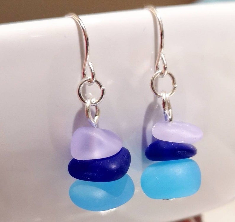 May include: Silver dangle earrings with three layered sea glass stones. The stones are light blue, dark blue, and lavender.