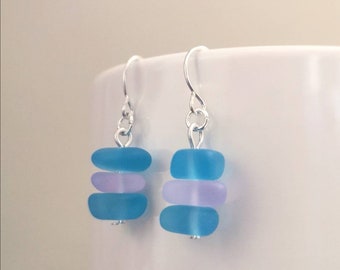Puede incluir: Un par de pendientes de plata con tres cuentas de vidrio de mar apiladas en tonos azules y lavanda.